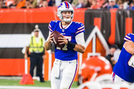 Quarterback Josh Allen stands in the pocket, ready to throw the football. He wears a white helmet (with a blue buffalo on it), blue jersey (number 17 written in white numbers), and white pants.