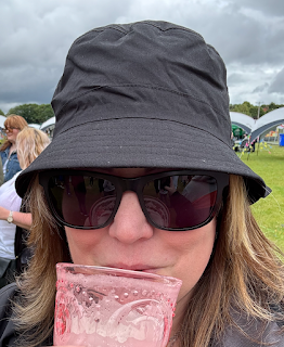 Me, a white woman with long brown hair, is standing in a field, wearing a black bucket hat and dark sunglasses, sipping a drink from a pink plastic glass.
