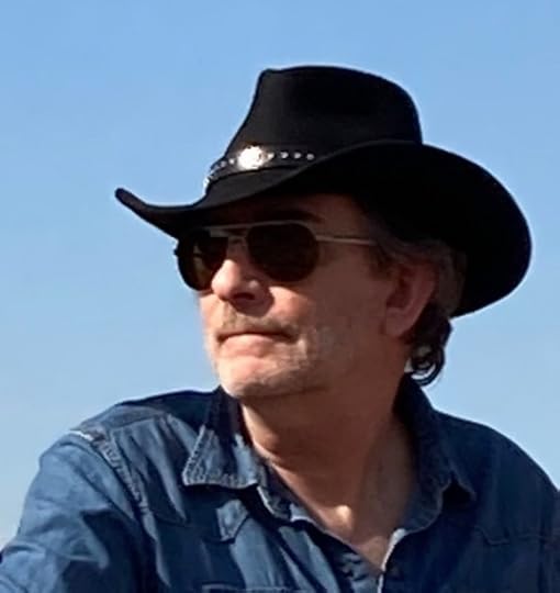 Jeffrey Mariotte is wearing a black had, sunglasses, and a blue denim shirt. There is a blue, cloudless sky behind him.