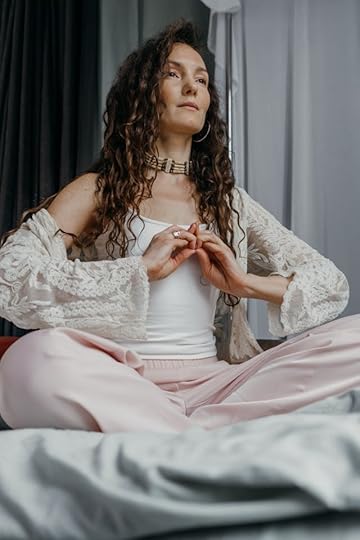 A woman with long curly hair sits cross-legged on a bed, forming a mudra gesture with her hands during a meditation session.