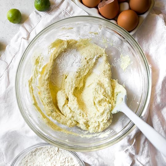 A mixing bowl with a spatula, containing key lime cookie dough batter at an intermediate stage of mixing. A small pile of baking powder, baking soda and salt rests on top of the dough.