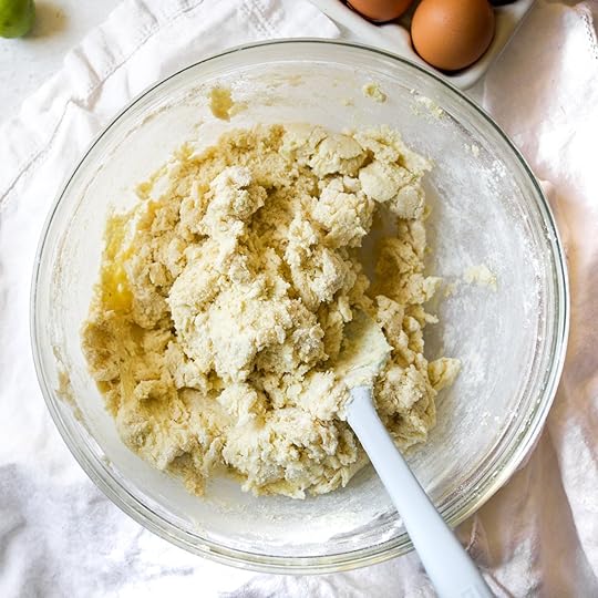 A glass bowl of fully mixed key lime cookie dough with a spatula, ready to be shaped. The dough is crumbly but cohesive, with visible flecks of lime zest.