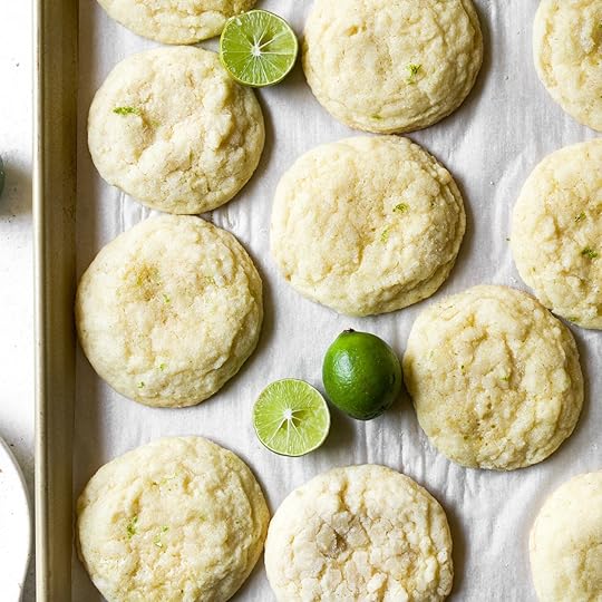 A baking sheet filled with freshly baked key lime cookies that have a crinkle top and are garnished with whole and halved fresh key limes.