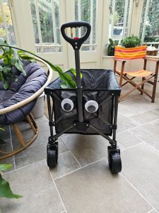 A festival trolley parked with the front towards us on a flagged stone floor. To the left part of a bamboo sofa is shown. To the right a pink,red, orange and green directors’s chair. Eyes are stuck on the trolley making it seem a little sad.