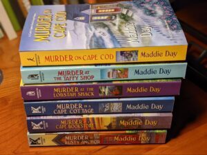 Stack of Six Cozy Capers Series books shown sideways with visible spines and titles.