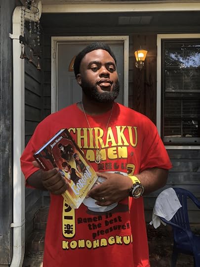 A Black man with facial hair and a red and yellow t-shirt holds a copy of his book in hardback outside his front door.