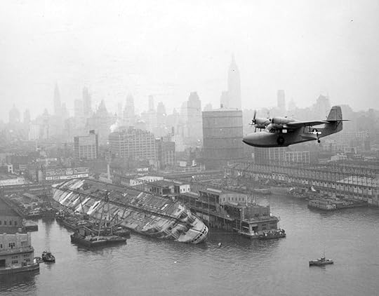 Black and white photograph of a Coast Guard plane flies over a ship that's listing severely to the left in the water.