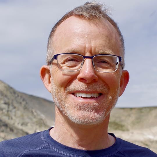 Sunlit head-and-shoulders of a smiling man with short hair and glasses, standing outside with a mountain ridge in the distance.