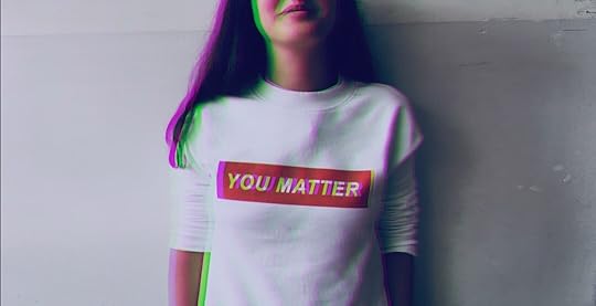 A person wearing a white long-sleeved shirt with the words “YOU MATTER” printed in bold yellow letters on a red rectangular background, standing against a plain wall.