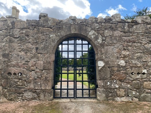gateway to Tolquhon Castle - old wall beneath blues sky, and an arched gate of black-painted metal bars.