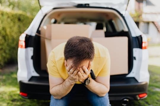 Stressed man sitting on the edge of a car trunk filled with cardboard boxes, covering his face with his hands in frustration or despair.