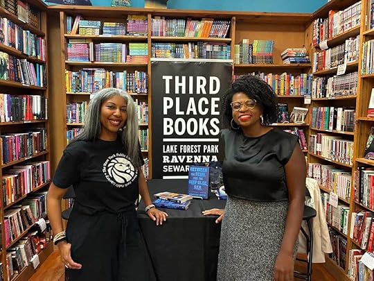 Khadijah in black (left) and Jodi-Ann Burry (right) in silk shirt and tweed skirt and glasses, both smiling in front of shelves of books and a display of Khadijah's book in the center