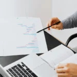 Person reviewing business data with charts on paper, using a pen to analyze trends, with a laptop open.