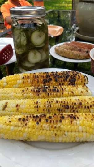 A plate of roasted corn on an outside table, with a jar of pickles and a plate of brats behind it.