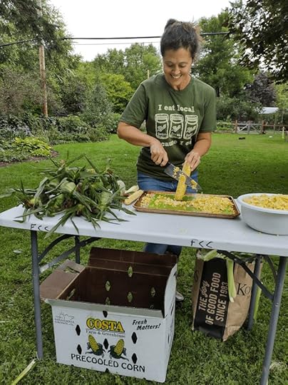A woman cutting corn off the cob with a knife into a pan. She's working outside on a table with corn husks on one side and a bowl of corn kernels on the other.