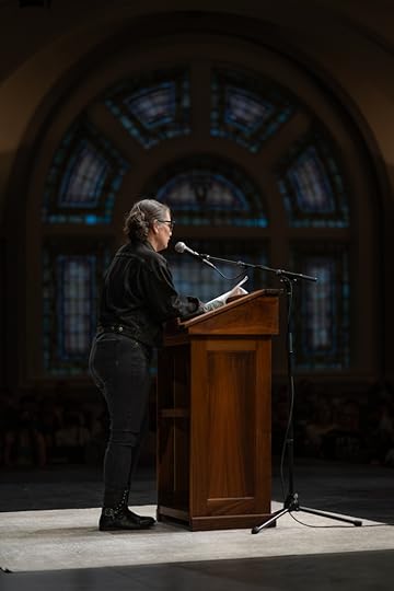 A writer in black denim at a lectern, framed by an arch of stained glass windows in the background