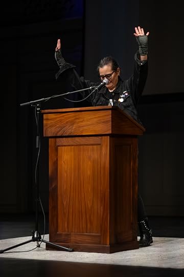 A dark-haired writer in black denim at a wooden lectern, arms raised, against a dark background
