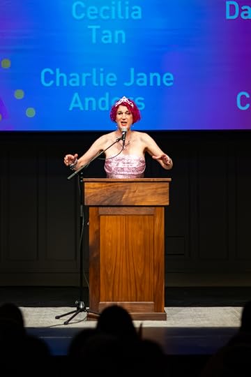An author with pink hair, in a pink corset dress and tiara, at a wooden lectern