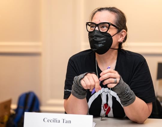 A small author with large glasses and a black KN94 mask, holding a pen, looking up at someone who is asking for an autograph