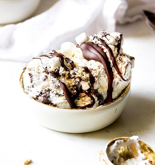 A close-up shot of a white bowl filled with creamy no-churn s'mores ice cream, drizzled with rich chocolate sauce and topped with crushed graham crackers and mini marshmallows. A gold ice cream scoop is visible next to the bowl.