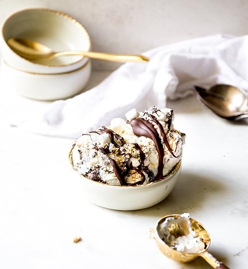A full bowl of no-churn s'mores ice cream, artistically drizzled with a rich chocolate sauce and sprinkled with crushed graham crackers and mini marshmallows. The image shows a full scene, including stacked bowls and an ice cream scoop.