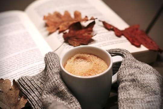 A white mug with a frothy drink is held by someone whose sweater sleeves are pulled over their hands. In the background is an open book with scattered autumn leaves.