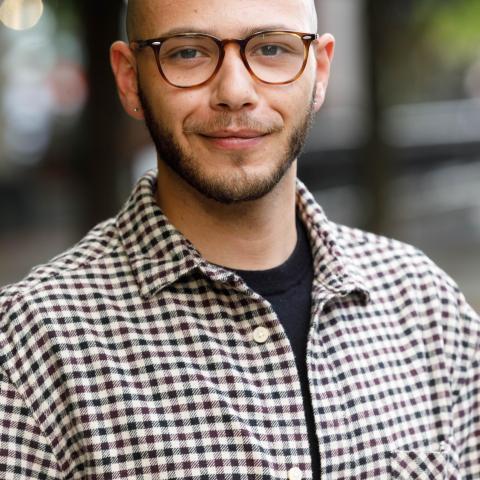 Tyler Coles, a person of African-American descent who has short facial hair and brown glasses, stands before a blurred cityscape. 