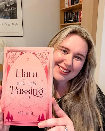 Author MG Hunt holding a copy of her short story Elara and the Passing near her face. She is in front of a book case and a cream wall.
