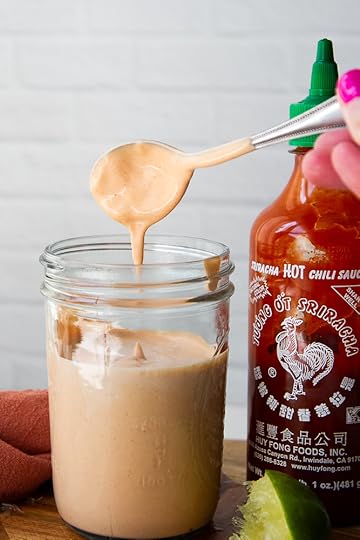 A mason jar with spicy mayo covered spoon hovering over the top with mayo dripping off. Sitting next to a bottle of sriracha sauce.