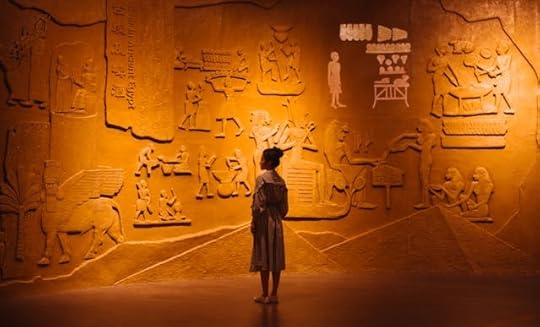 A young girl stands alone in front of an illuminated wall covered in ancient carvings and symbols in a museum exhibit.