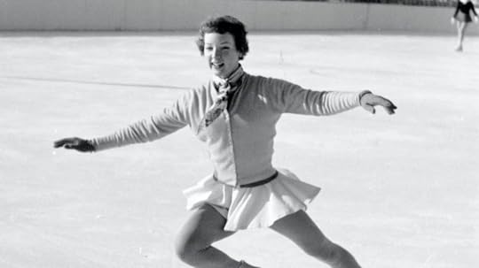 A female figure skater with short hair smiles and skates outdoors, arms outstretched. Another blurred skater is in the background.