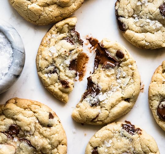 Close-up of a chewy almond chocolate chip cookie split in half with melted chocolate and flaky sea salt, surrounded by other cookies on a white marble surface next to a small bowl of salt.