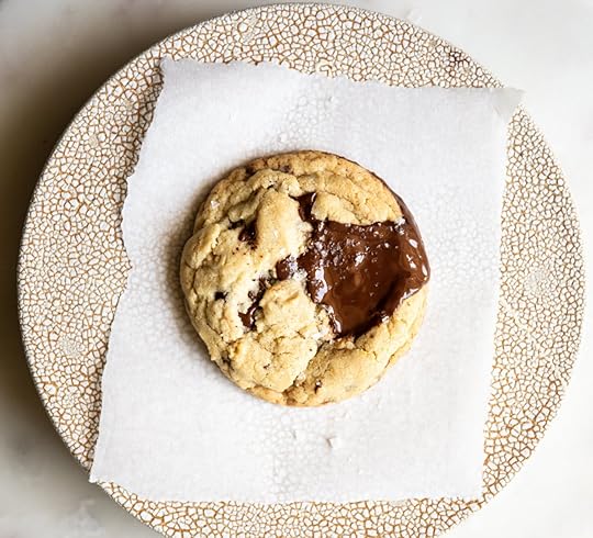 A single, warm almond chocolate chip cookie with melted chocolate pooled on top, resting on a small square of parchment paper on a decorative cracked-glaze plate.