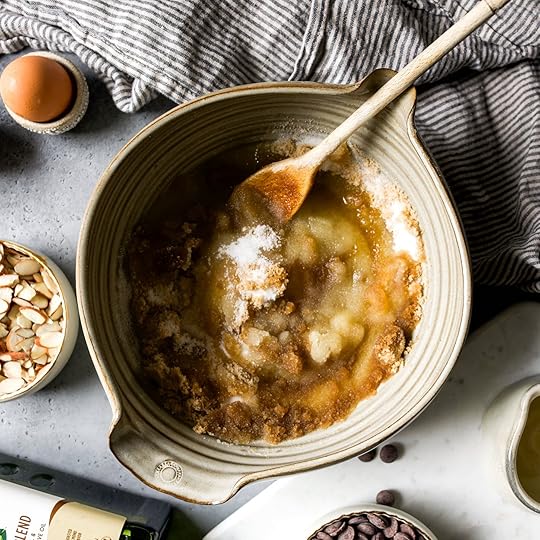Overhead view of a ceramic mixing bowl with wet and dry ingredients, including brown sugar, white sugar, and melted butter, being stirred with a wooden spoon, surrounded by an egg, sliced almonds, chocolate chips, and olive oil.