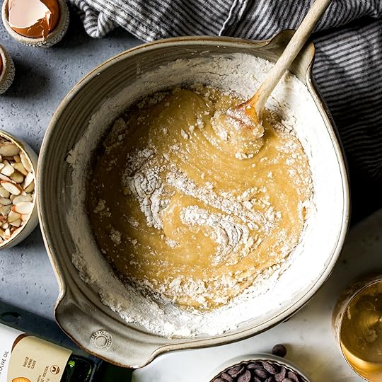A wooden spoon mixes flour into a creamy batter in a large ceramic bowl, surrounded by ingredients like sliced almonds, chocolate chips, and a cracked egg.