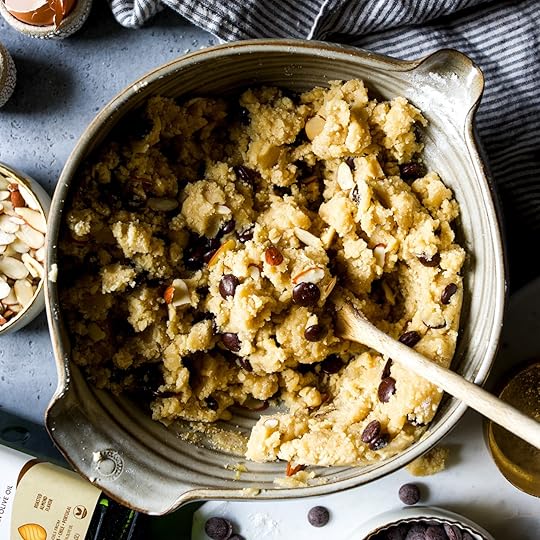 Fully mixed almond chocolate chip cookie dough, with chocolate chips and sliced almonds evenly distributed, is shown in a ceramic mixing bowl with a wooden spoon.