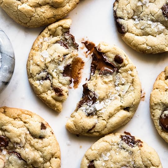 A freshly baked almond chocolate chip cookie, broken in half to reveal gooey melted chocolate inside, sprinkled with flaky sea salt, surrounded by other cookies on a light marble surface.