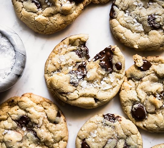 A close-up overhead view of golden-brown almond chocolate chip cookies, one broken open to reveal melted chocolate and a sprinkle of flaky sea salt, on a white surface with a small gray salt cellar.