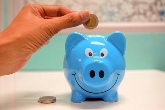 A hand placing a coin into a smiling blue piggy bank, symbolizing financial freedom affirmations and joyful money habits