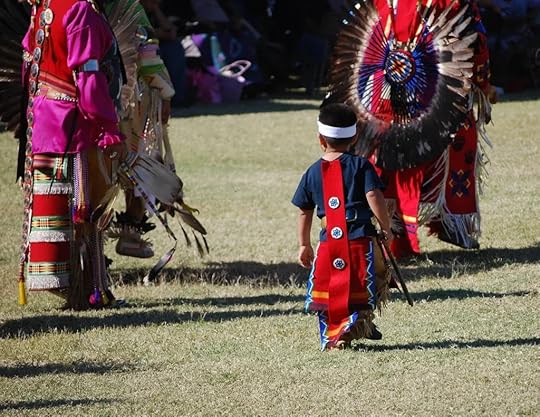 A young Native American boy dressed in traditional clothing, including a headdress, standing on a grassy field.