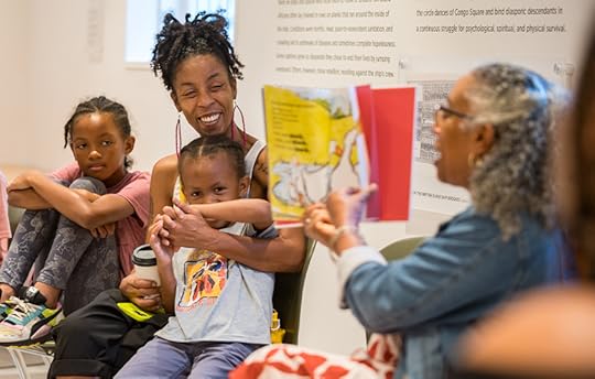 A woman holds up a copy of a colorful book as she reads to a small group of mothers and children.