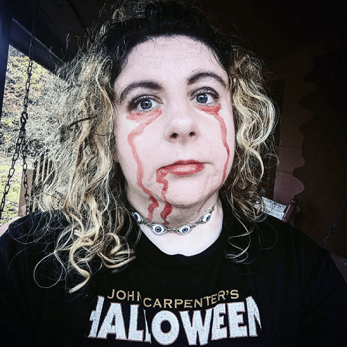 A close-up photo of the author standing outside. A white person with dyed blond hair wears a black shirt with JOHN CARPENTER'S HALLOWEEN visible, a choker with several blue eyes, and fake blood that runs from their eyes.