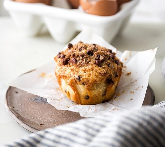 A close-up shot of a single bakery-style chocolate chip muffin with a tall crown, a cinnamon brown sugar streusel topping, and melty chocolate chips, resting on a small plate.