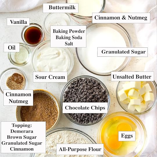 Overhead shot of all the ingredients for a chocolate chip muffin recipe laid out on a white counter. The ingredients include all-purpose flour, butter, eggs, buttermilk, sour cream, baking soda, baking powder, salt, vanilla, oil, chocolate chips, and a cinnamon brown sugar streusel topping.