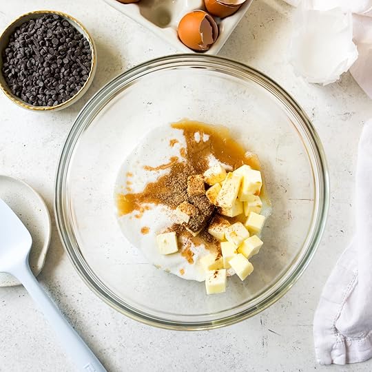 An overhead shot of a clear mixing bowl with cubed butter, granulated sugar, oil, vanilla, cinnamon, and nutmeg. In the background, there is a small bowl of chocolate chips and a carton of cracked eggs.