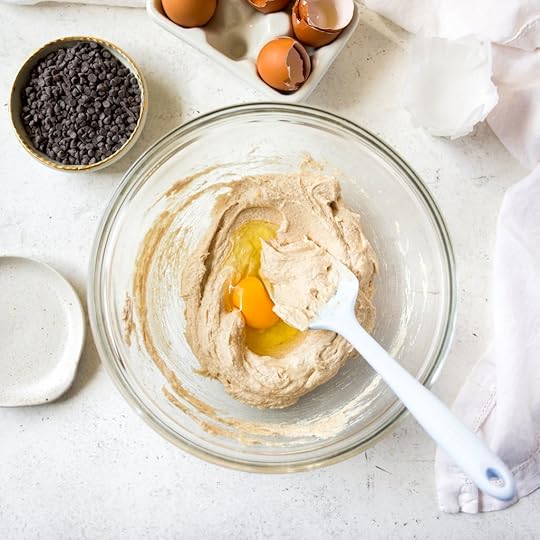 An overhead shot of a clear mixing bowl with creamed butter and sugar, with an egg cracked in the center. A white spatula is resting in the bowl.