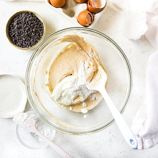 An overhead shot of a clear mixing bowl containing a creamed butter and sugar mixture. A white spatula, sour cream, and a spoonful of baking powder, baking soda and salt are being added to the bowl.