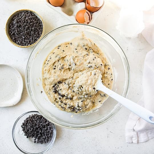 An overhead shot of a clear mixing bowl filled with thick chocolate chip muffin batter. A white spatula rests in the bowl and bowls of mini chocolate chips are visible in the background.
