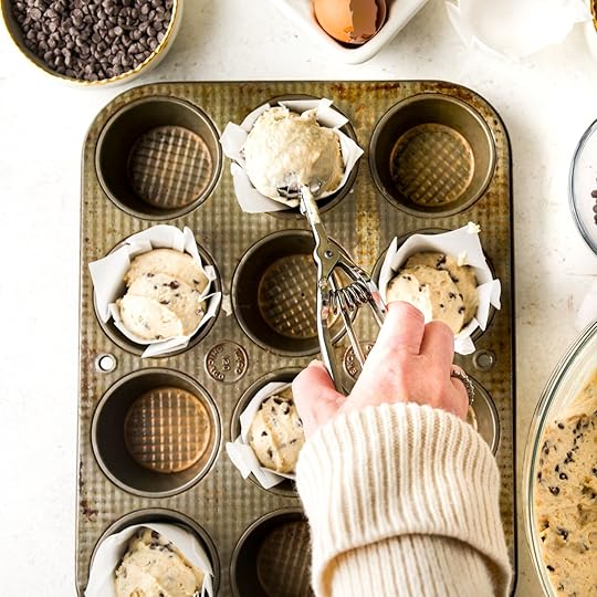 An overhead shot of hands using an ice cream scoop to fill a muffin tin, lined with white paper liners, with chocolate chip muffin batter.