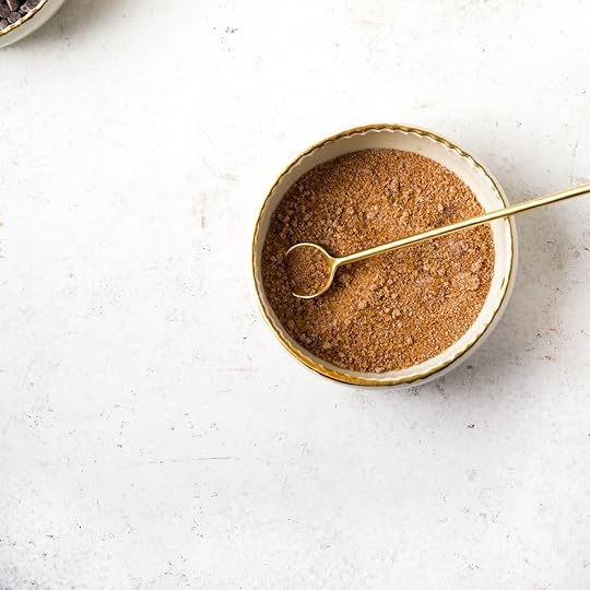 A small white bowl with a gold rim containing a mixture of brown sugar, Demerara sugar, granulated sugar, and cinnamon for a streusel topping, with a gold spoon resting inside.